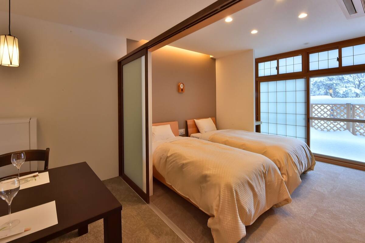 A hotel room with two beds, a dining table with a wine glass, a pendant light, and a view of a snowy deck through large windows.