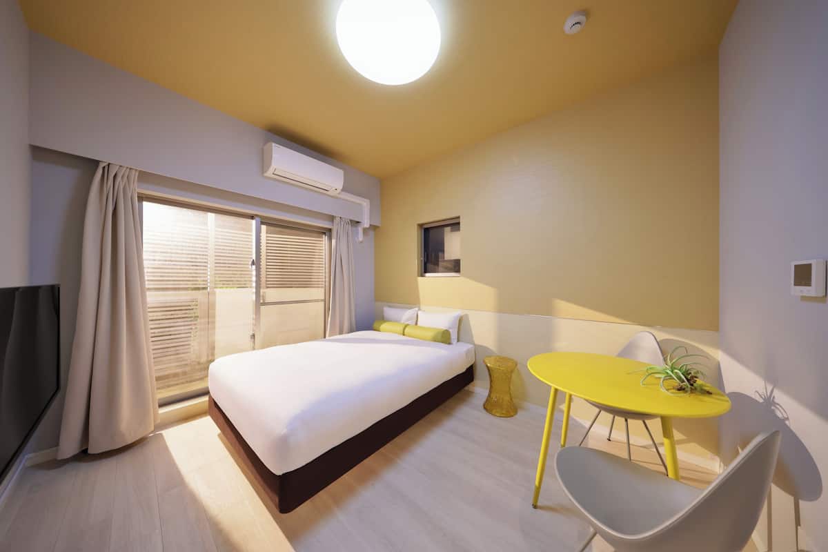 A hotel room with a bed, a small table with a plant, a chair, and a window with curtains.
