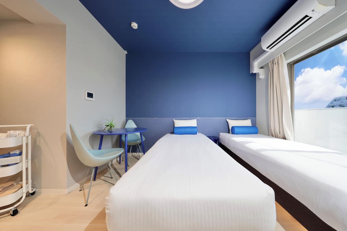 A hotel room with a bed, a desk, a chair, a small table, and an air conditioning unit.