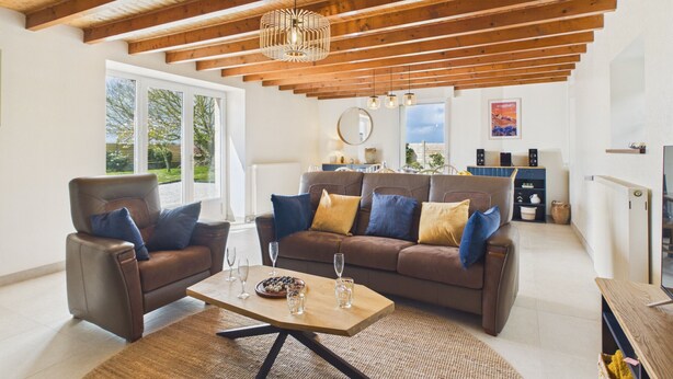 Amplia sala de estar con techos altos, vigas de madera, un sofá de cuero marrón con cojines, una mesa de centro y una zona de comedor al fondo.