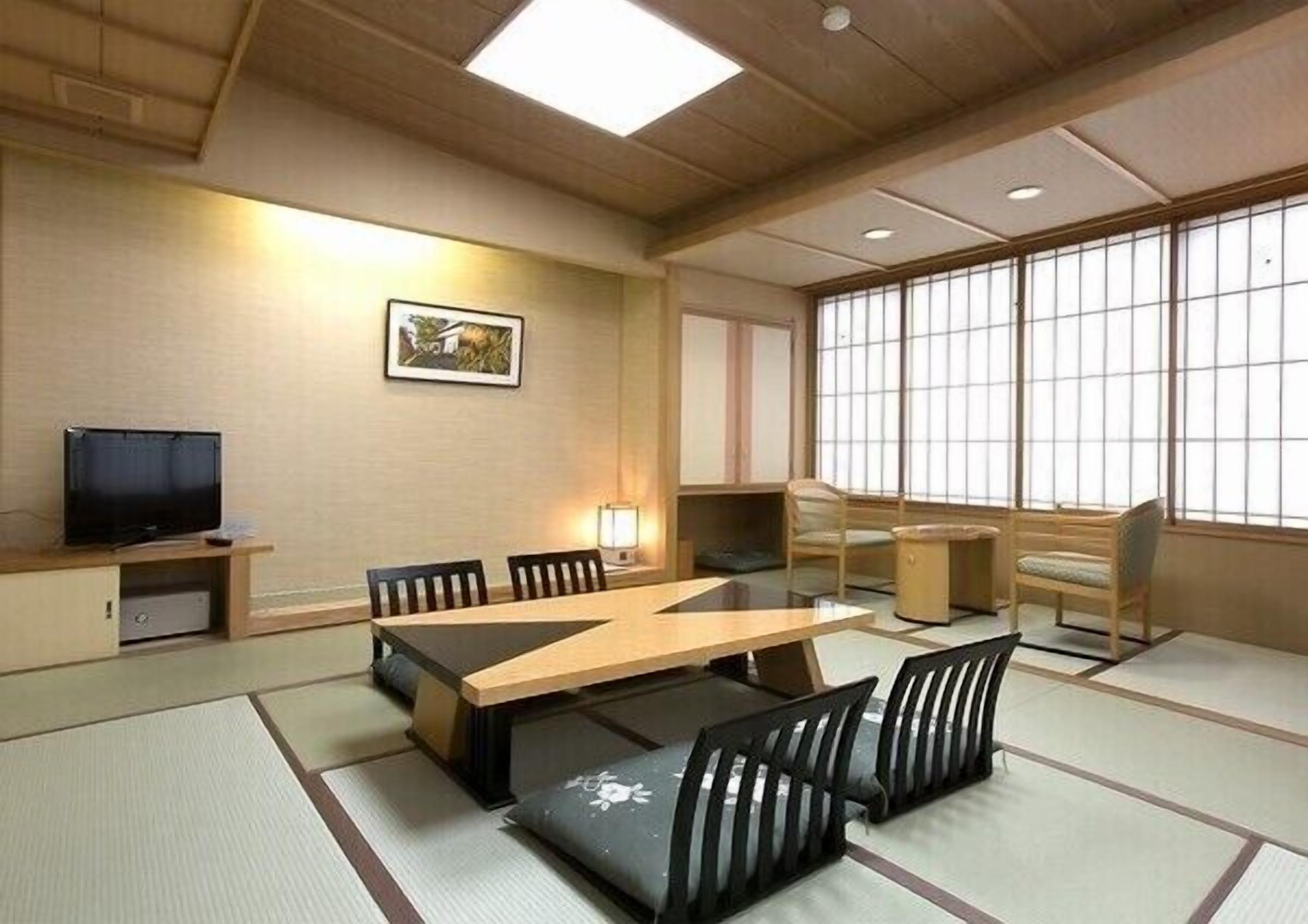 A traditional Japanese-style room with tatami flooring, a low table, chairs, a television, and large windows.