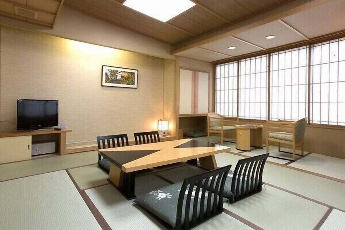 A traditional Japanese-style room with tatami flooring, a low table, chairs, a television, and large windows.
