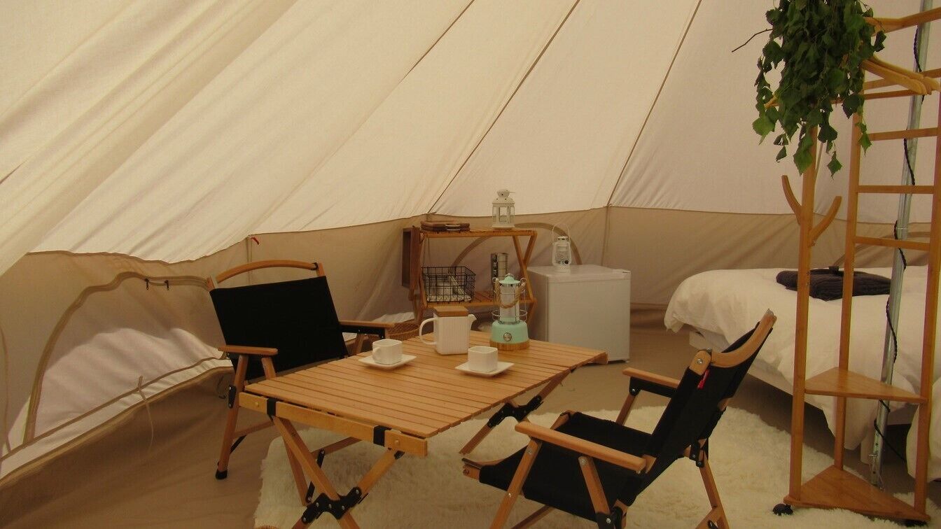 Uma tenda com uma mesa de madeira, cadeiras, uma pequena estante com objetos e uma planta pendurada.