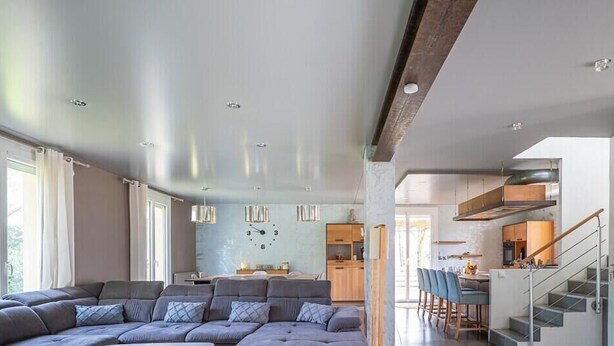 Una sala moderna con un sofá seccional grande, un comedor y una cocina al fondo, y una escalera que sube a un nivel superior.