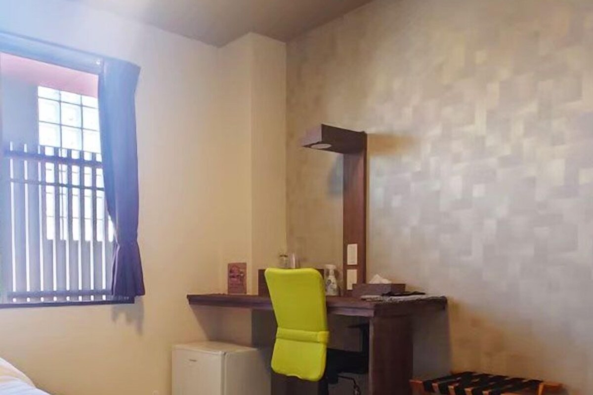 A hotel room with a bed, a desk with a chair, a small refrigerator, and a window with curtains.