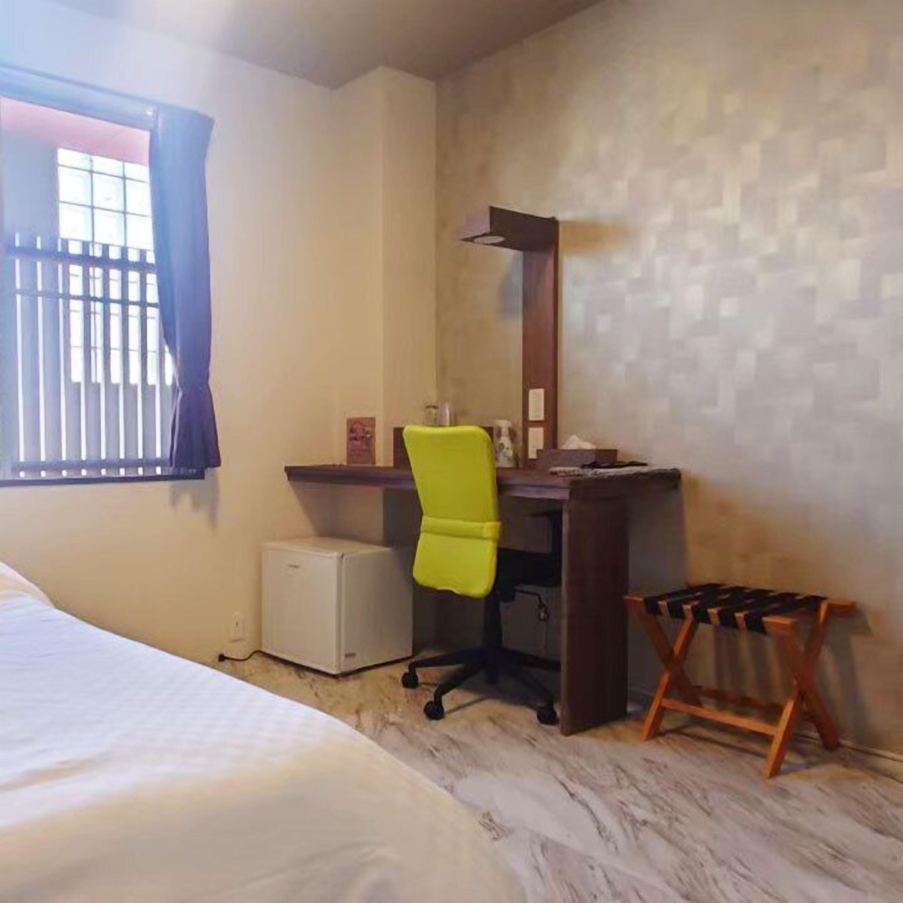 Une chambre d’hôtel comprenant un lit, un bureau avec une chaise, un petit réfrigérateur et une fenêtre avec des rideaux.