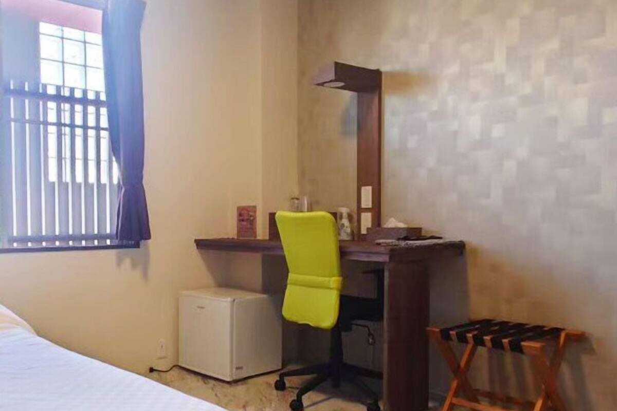 Une chambre d’hôtel comprenant un lit, un bureau avec une chaise, un petit réfrigérateur et une fenêtre avec des rideaux.