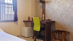 Une chambre d’hôtel comprenant un lit, un bureau avec une chaise, un petit réfrigérateur et une fenêtre avec des stores.