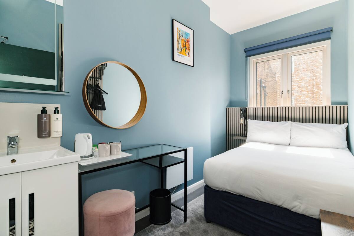 A bedroom with a bed, a desk, a mirror, a window, and a bathroom area with a sink and toiletries.