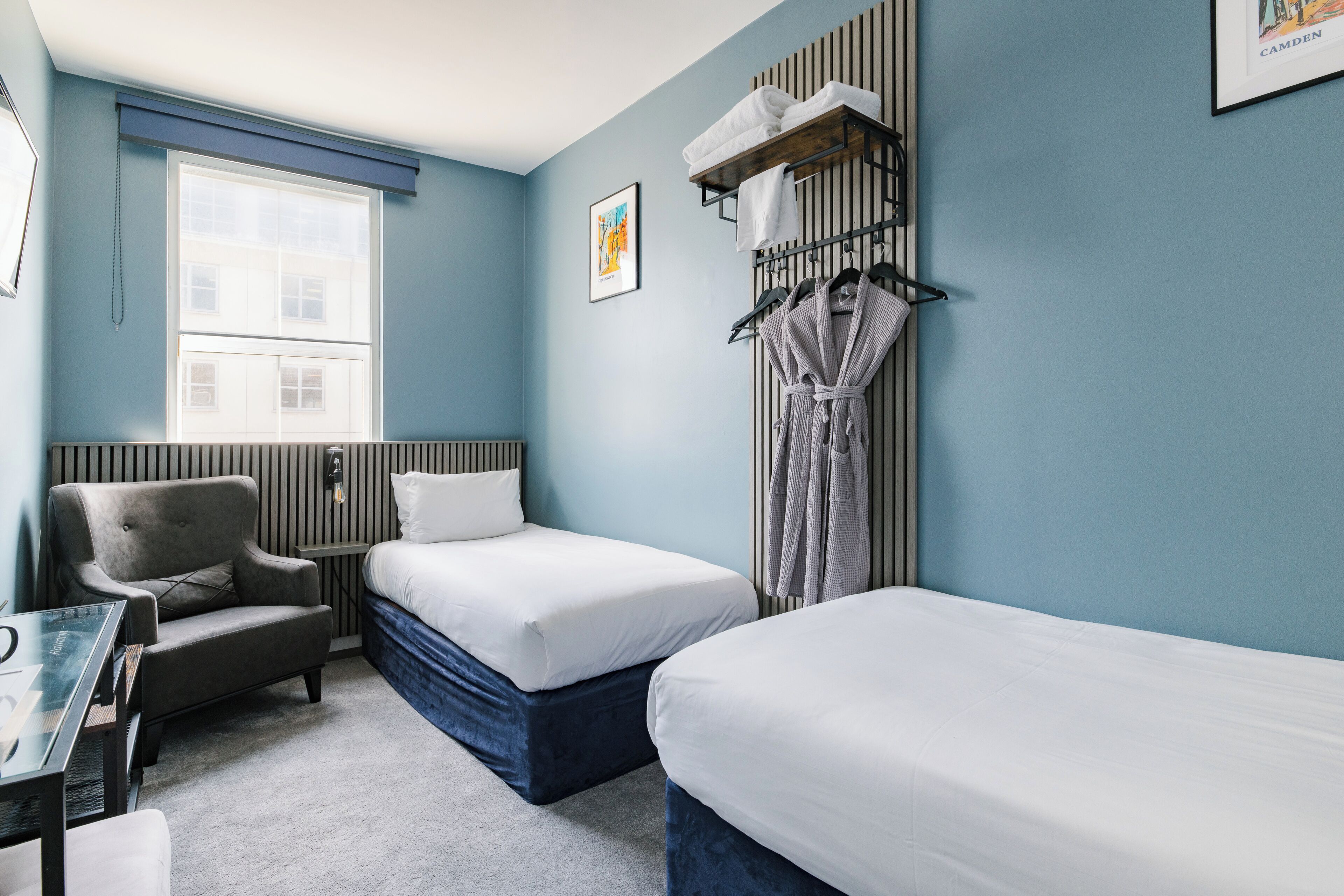 A hotel room with two beds, a chair, a table, a window, and a towel rack.