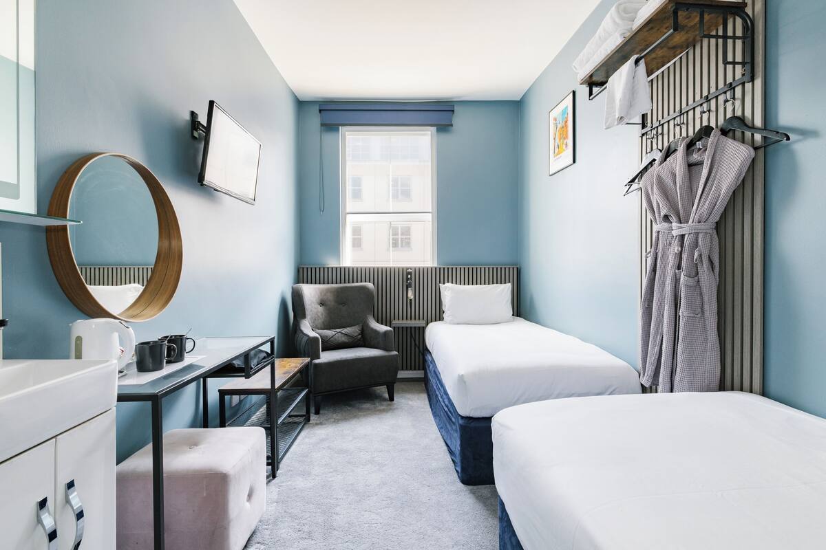 A hotel room with a bed, a chair, a desk, a mirror, a window, and a robe rack.