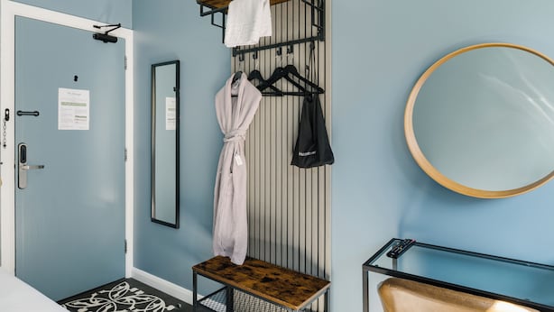 A hotel room with a wooden bench, a mirror, a coat rack with towels and a robe, and a door with a sign.