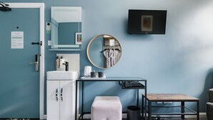 A bathroom with a sink, mirror, and toilet, a round mirror on the wall, a black trash bin, and a small wooden table.