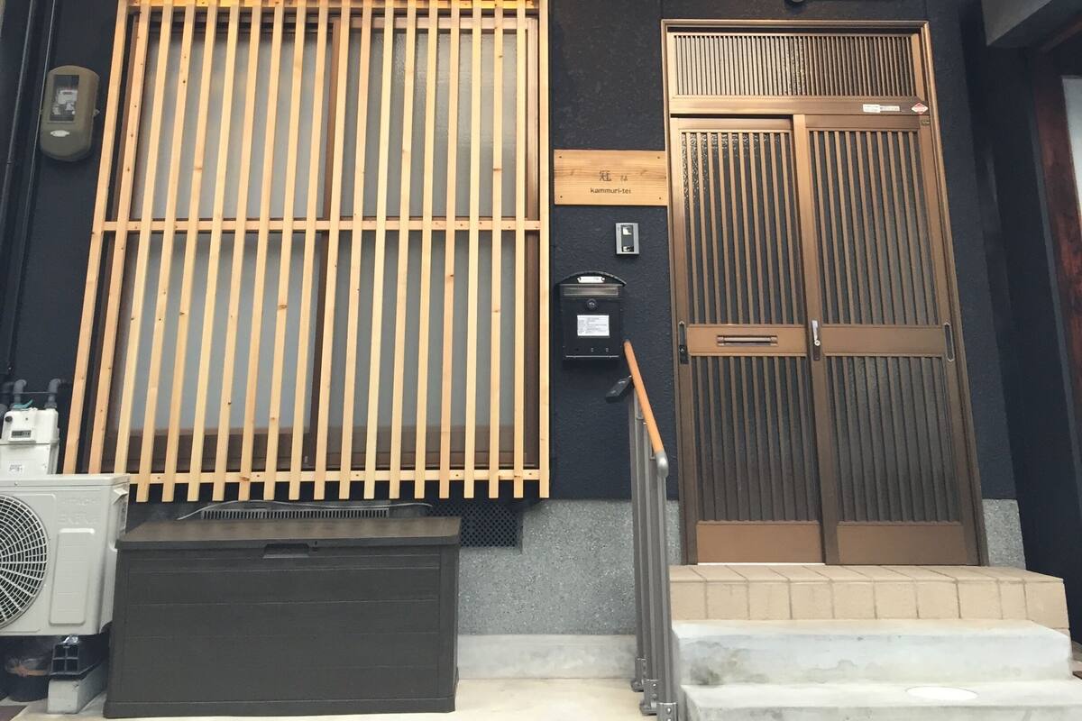 A two-story building with a wooden balcony, a black door with vertical slats, and a small air conditioning unit on the ground floor.
