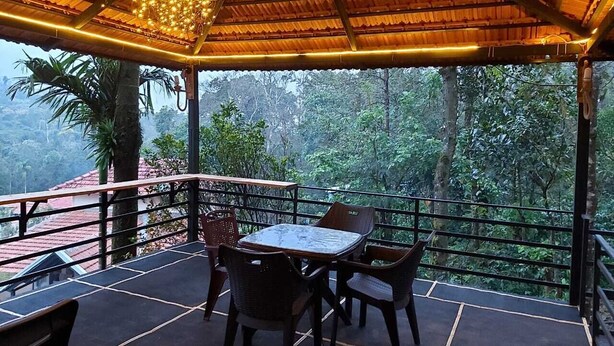 A covered outdoor seating area with a table and chairs, surrounded by a railing, overlooking a forested landscape.