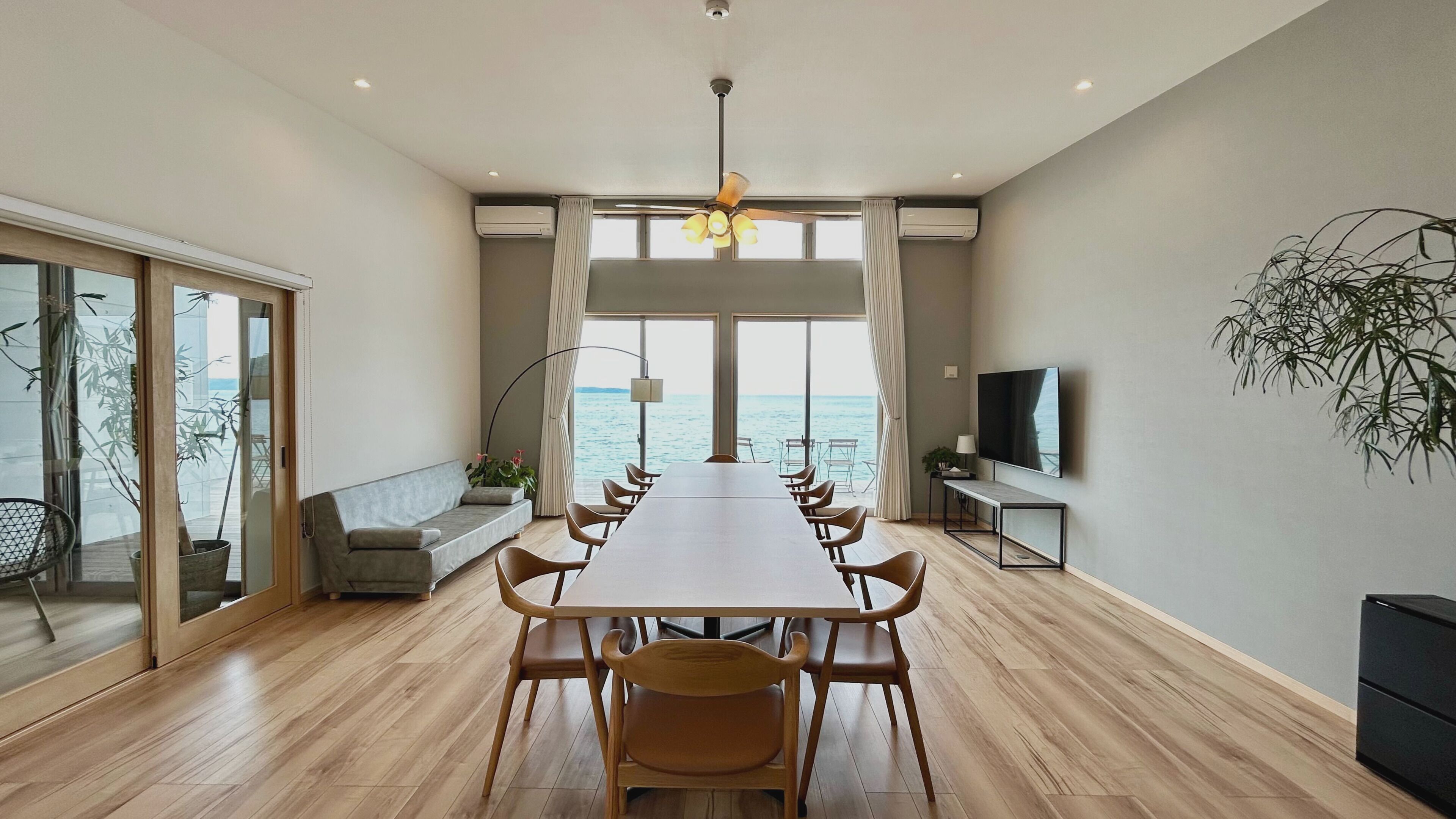 Une salle à manger moderne avec une longue table, des chaises, un canapé, un téléviseur et une vue sur la mer.