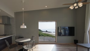 A modern kitchen with a dining area, a ceiling fan, and a large window with a view of the beach. - Alfacio Resort Stay Itoshima (Shimukappu)