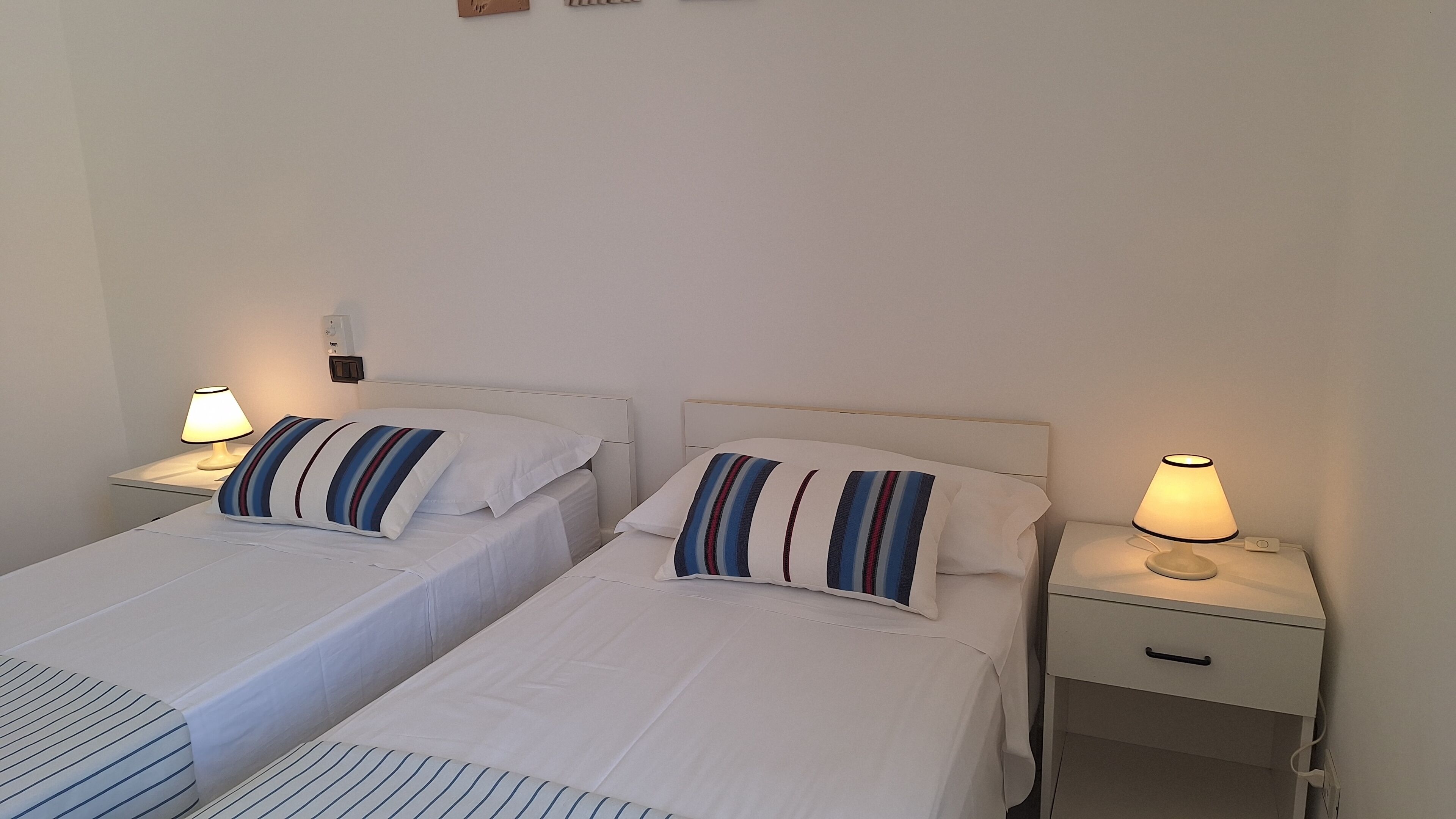 A hotel room with two single beds, each with a striped pillow and a bedside lamp. There is a small drawer on the right side of the bed.