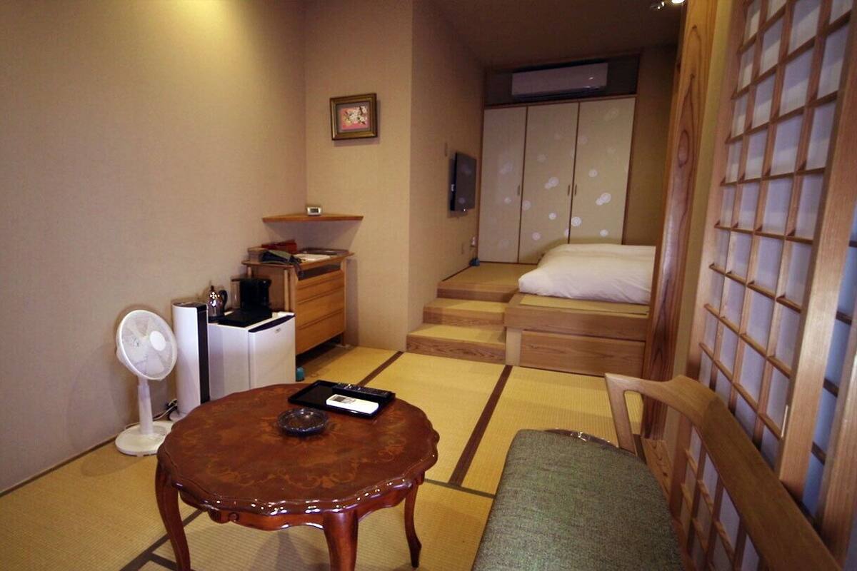 A traditional Japanese room with tatami flooring, a low wooden table, a chair, a bed with a wooden headboard, and a wardrobe.
