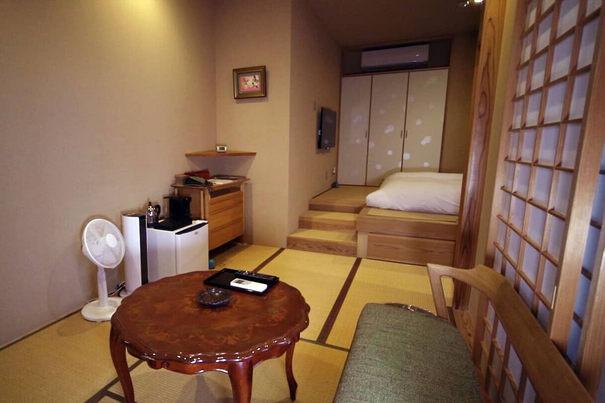Une pièce de style japonais traditionnel, avec un sol recouvert de tatamis, une petite table en bois, une chaise, un lit doté d’une tête de lit en bois et une armoire.