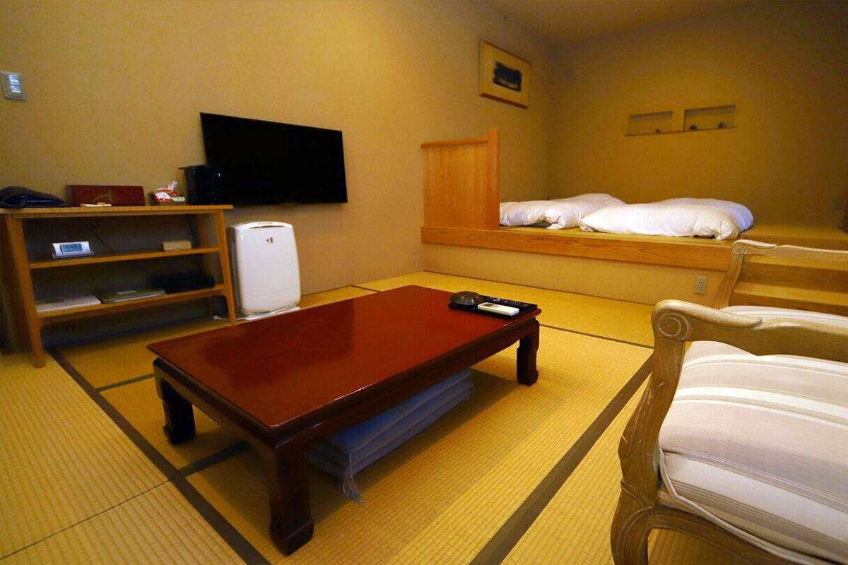 A room with tatami flooring, a wooden coffee table, a television, a bed with white bedding, a chair, and a shelf with items.