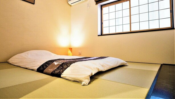 A room with tatami flooring, a bed with a patterned blanket, a window with grid panes, and an air conditioning unit. - Kyorana Kyogetsutei (Kyoto)