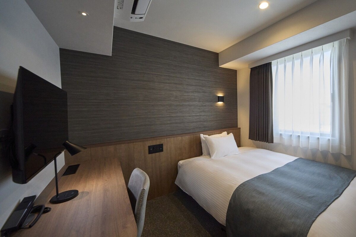 Habitación de hotel con cama, escritorio, silla, televisor y ventana con cortinas.