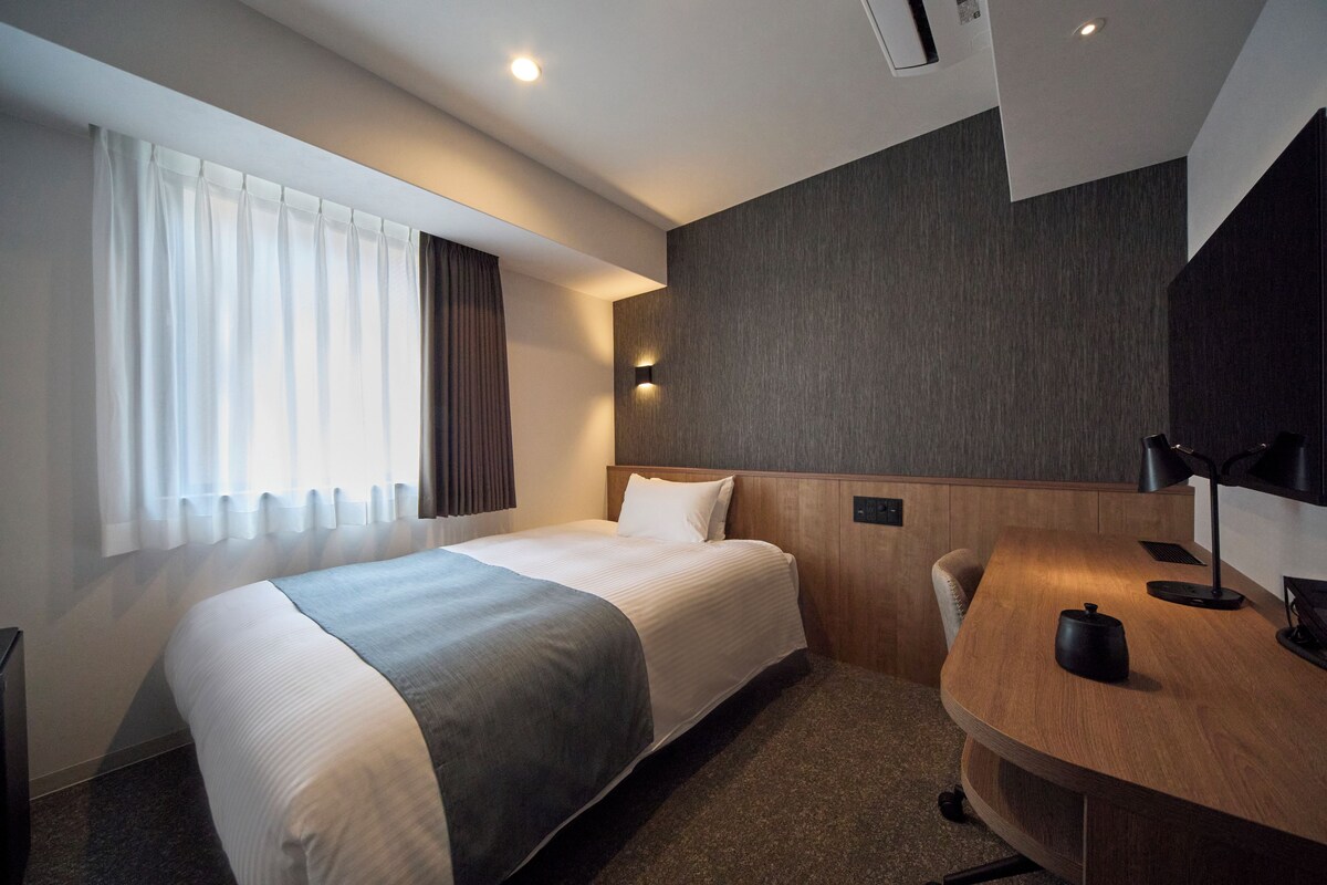 Una habitación de hotel con cama, escritorio, silla, lámpara y ventana con cortinas.