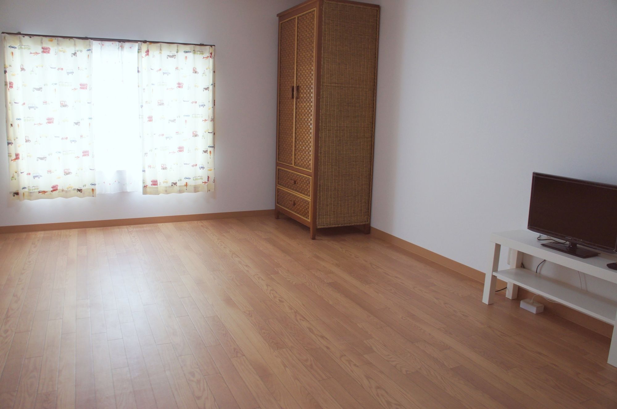 Una habitación con piso de madera, un armario alto, un televisor sobre un soporte blanco y una ventana con cortinas estampadas.