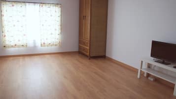 Una habitación con piso de madera, un armario alto, un televisor sobre un soporte blanco y una ventana con cortinas estampadas.