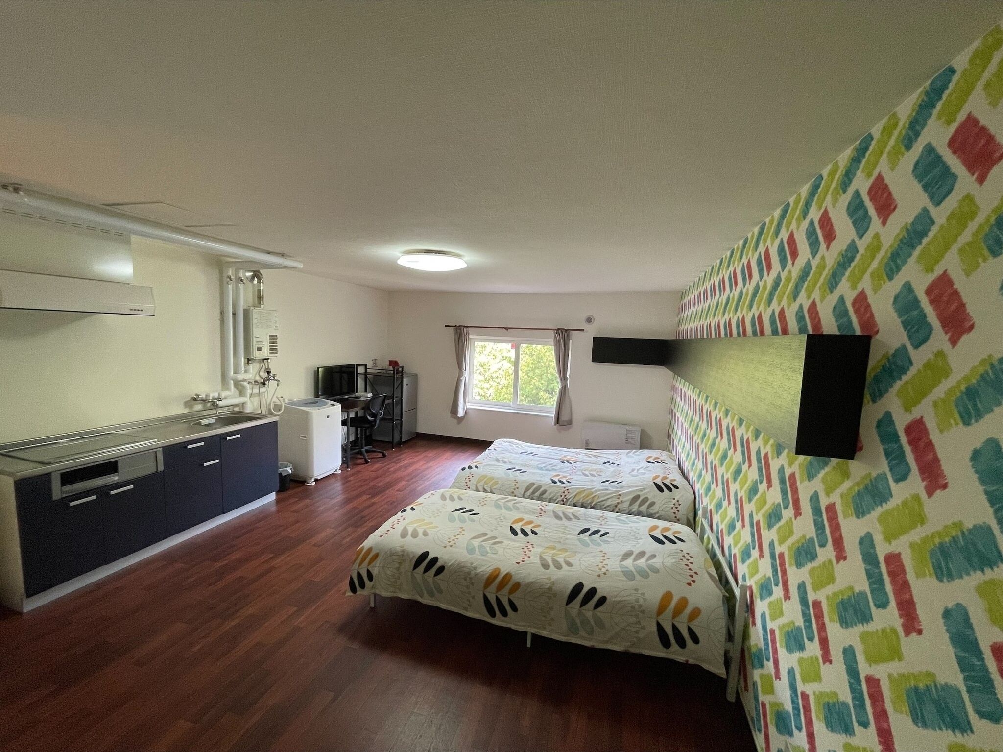 A room with a bed, a desk, a kitchenette, and a window.
