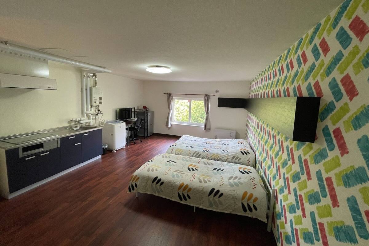 A room with a bed, a desk, a kitchenette, and a window.