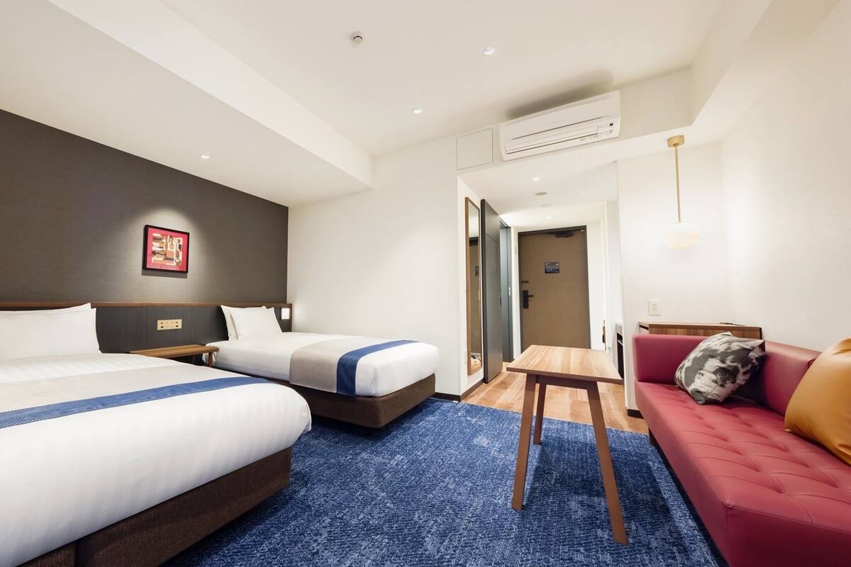 A hotel room with two beds, a small table, a sofa, and an air conditioning unit.