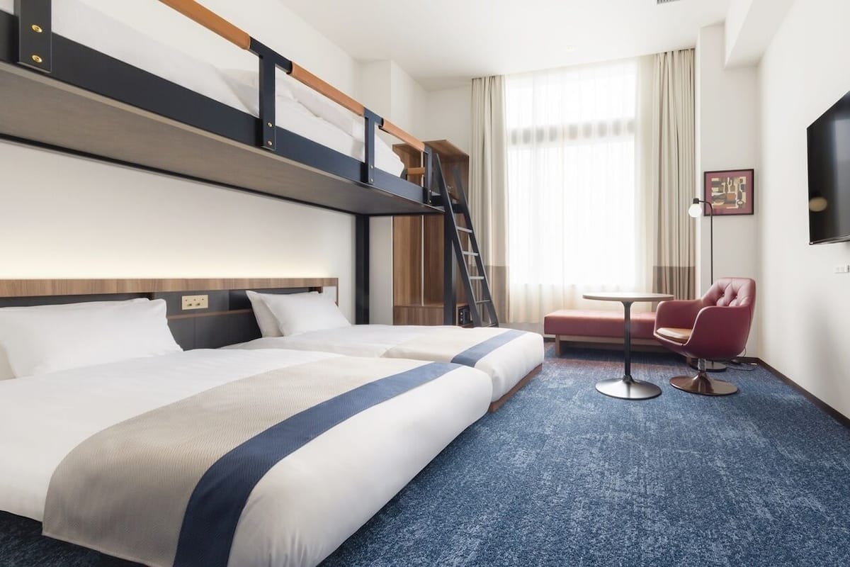 A hotel room with a bunk bed, a desk, a chair, a TV, and a window with curtains.