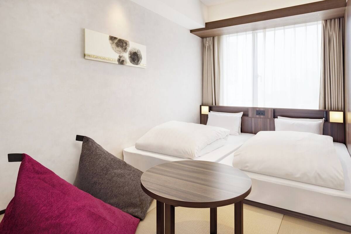 A hotel room with a bed, a small table, a chair, a painting on the wall, and a window with curtains.