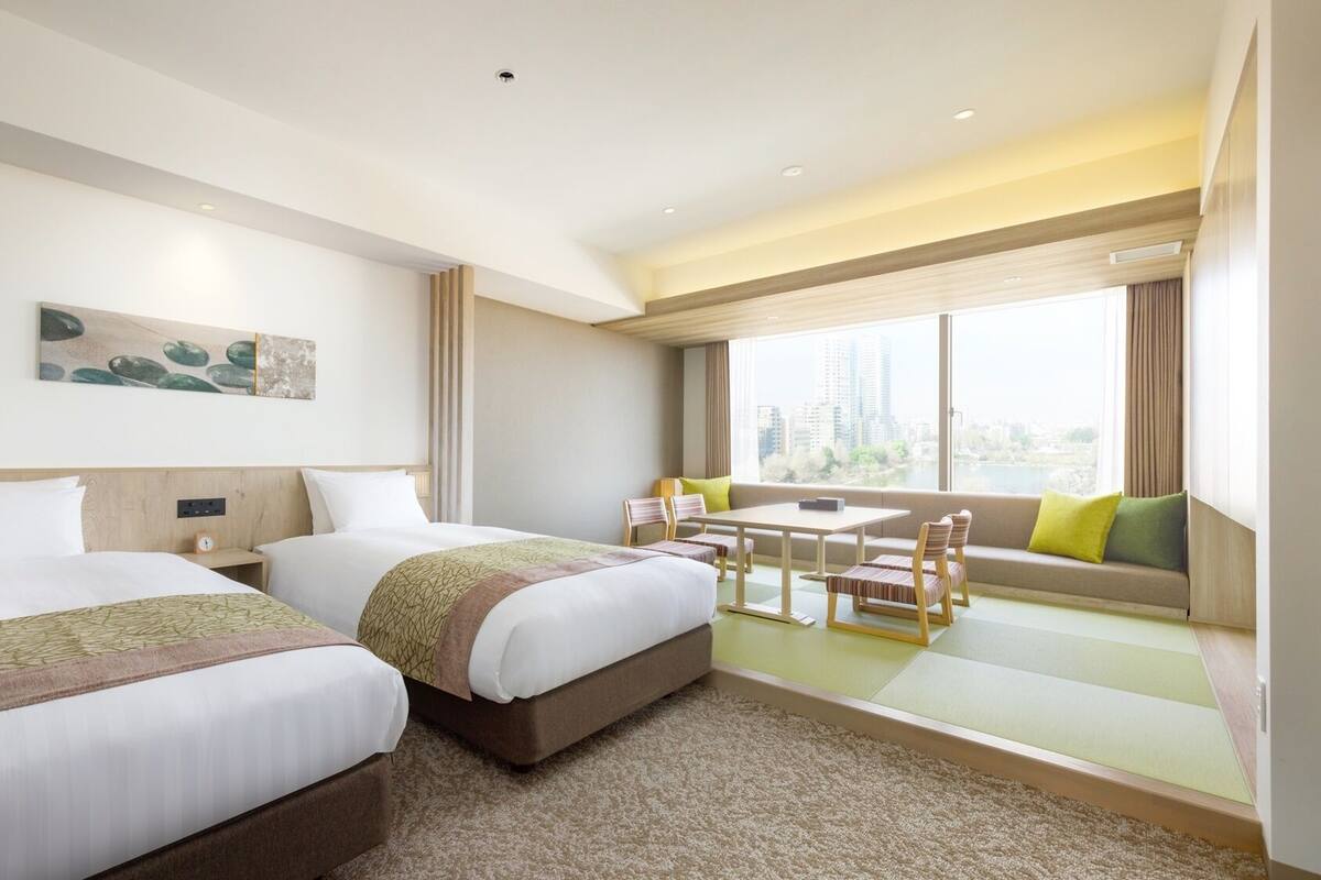 A hotel room with two beds, a dining table, chairs, a sofa, and a large window with a city view.