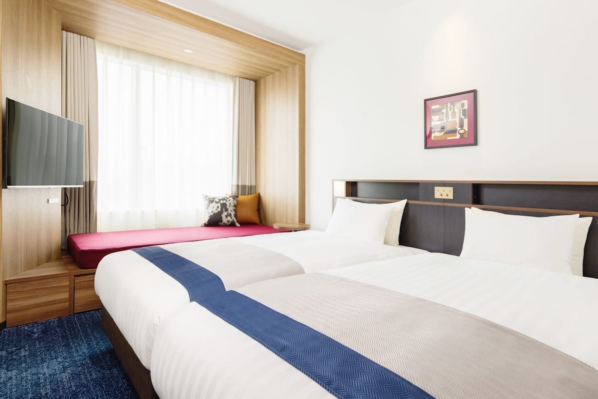 A hotel room with two beds, a TV, a window with curtains, a bedside table, and a wall-mounted artwork.