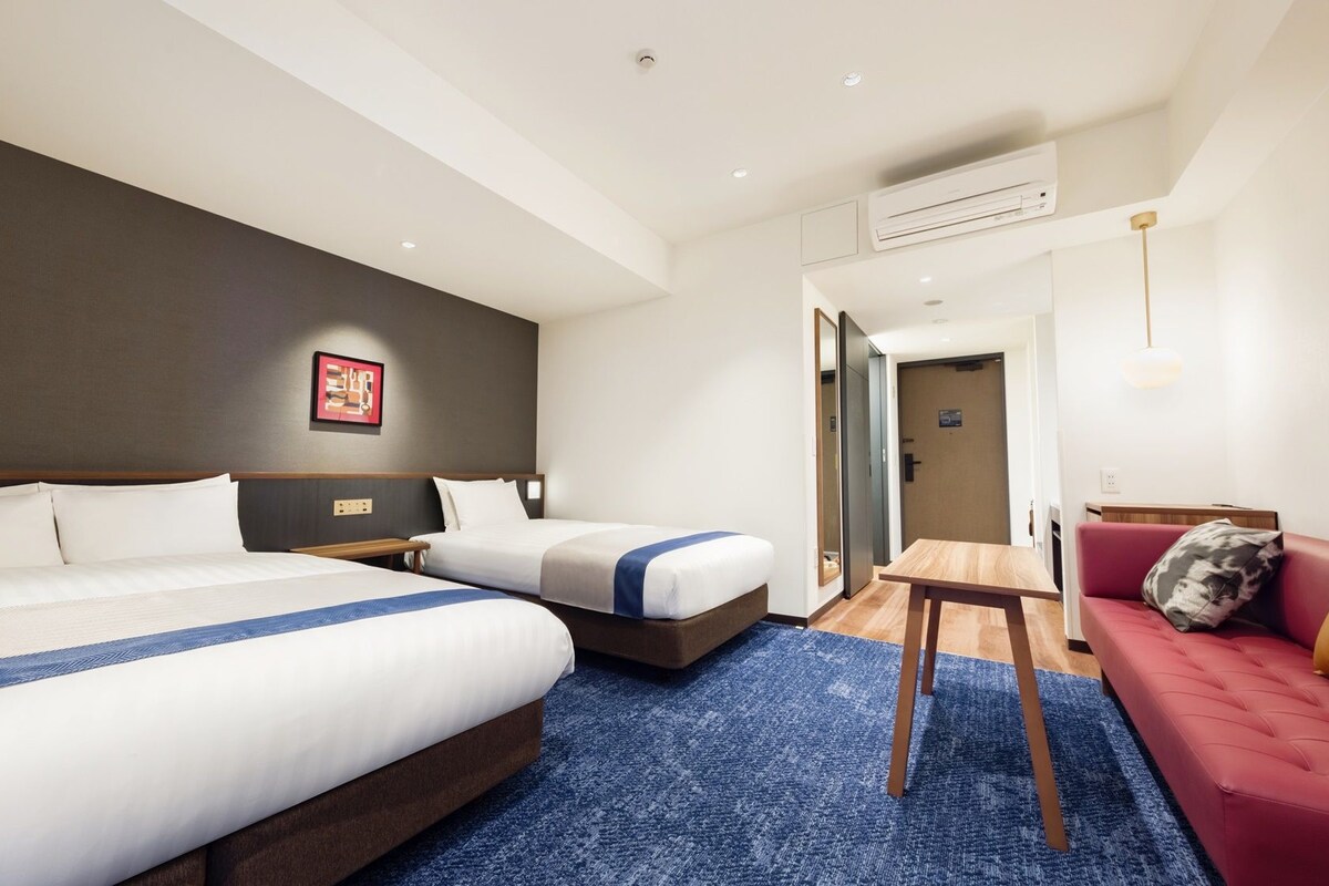 A hotel room with two beds, a small table, a sofa, and an air conditioning unit.