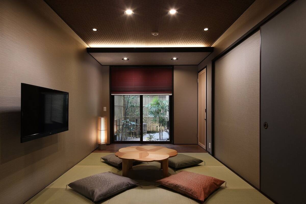 A traditional Japanese room with tatami flooring, a low table, cushions, a sliding door, and a television.
