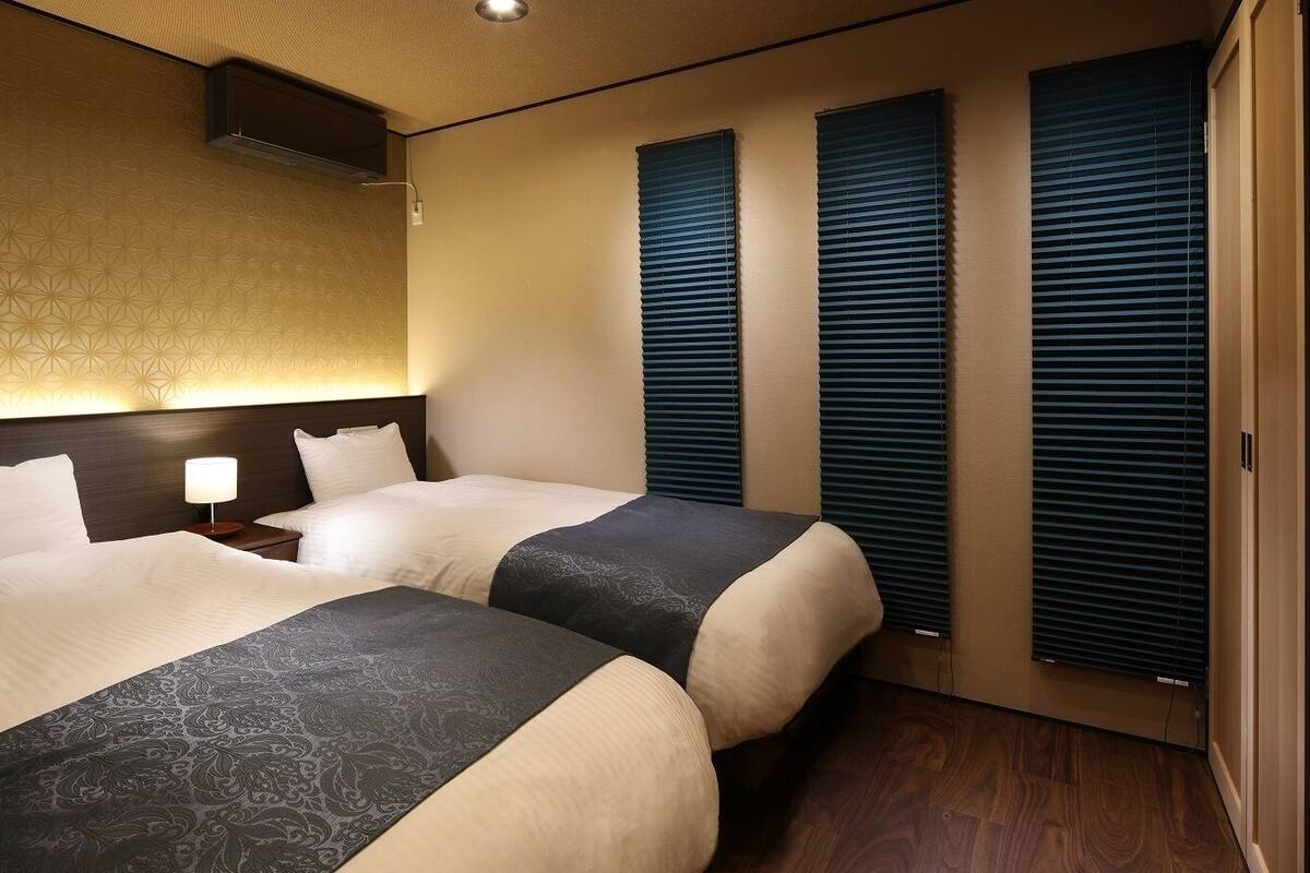 A hotel room with two beds, a headboard with a light fixture, a bedside table with a lamp, and a window with blinds.