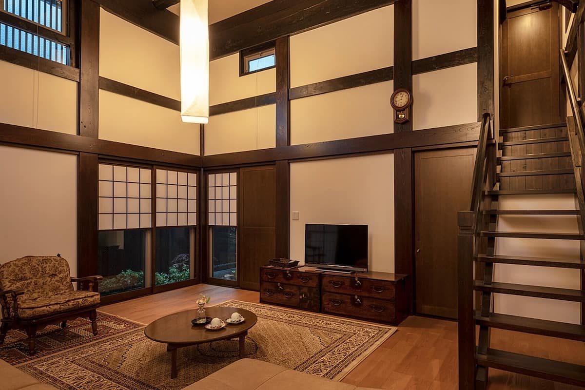 Una habitación tradicional japonesa con suelo de tatami, una mesa de madera baja, un sofá, un televisor y una escalera.