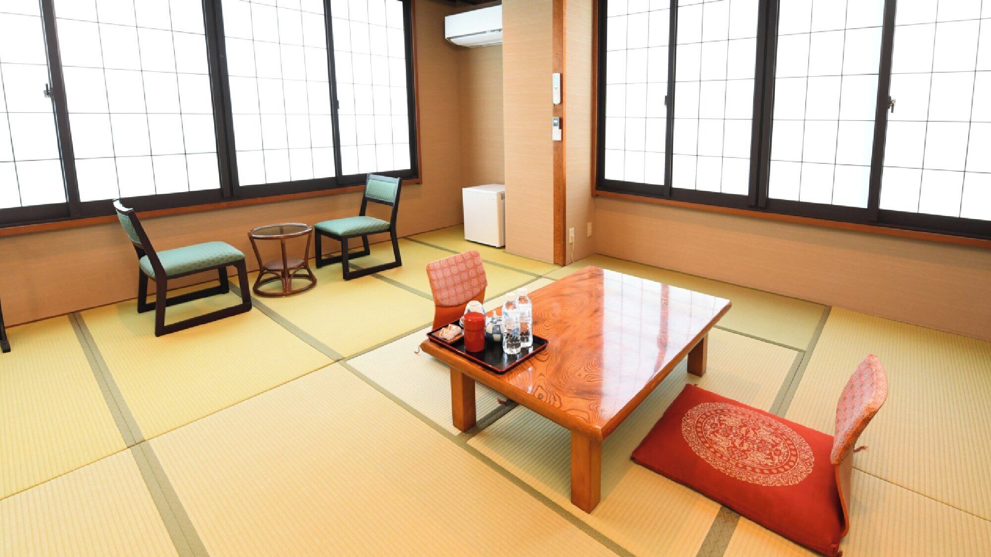 Une pièce avec un sol recouvert de tatamis, une table en bois, deux chaises, une petite table ronde et un coussin rouge.