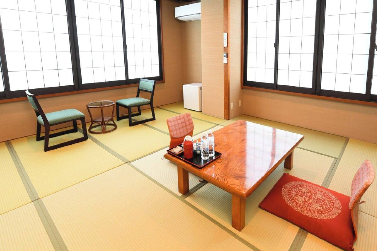 A tatami mat room with a wooden table, two chairs, a small round table, and a red cushion.