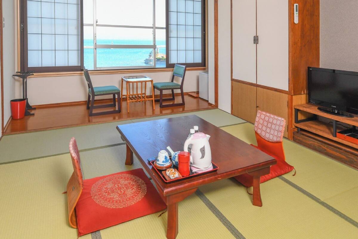 A traditional Japanese room with tatami flooring, a low table with tea set, a television, and a view of the sea.