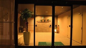 Entrada de hotel con puerta corrediza, planta en maceta, pared de ladrillo y mostrador de recepción.