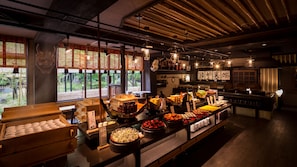 Um restaurante com diversos alimentos expostos no balcão, mobiliário de madeira e grandes janelas que permitem entrada de luz natural.