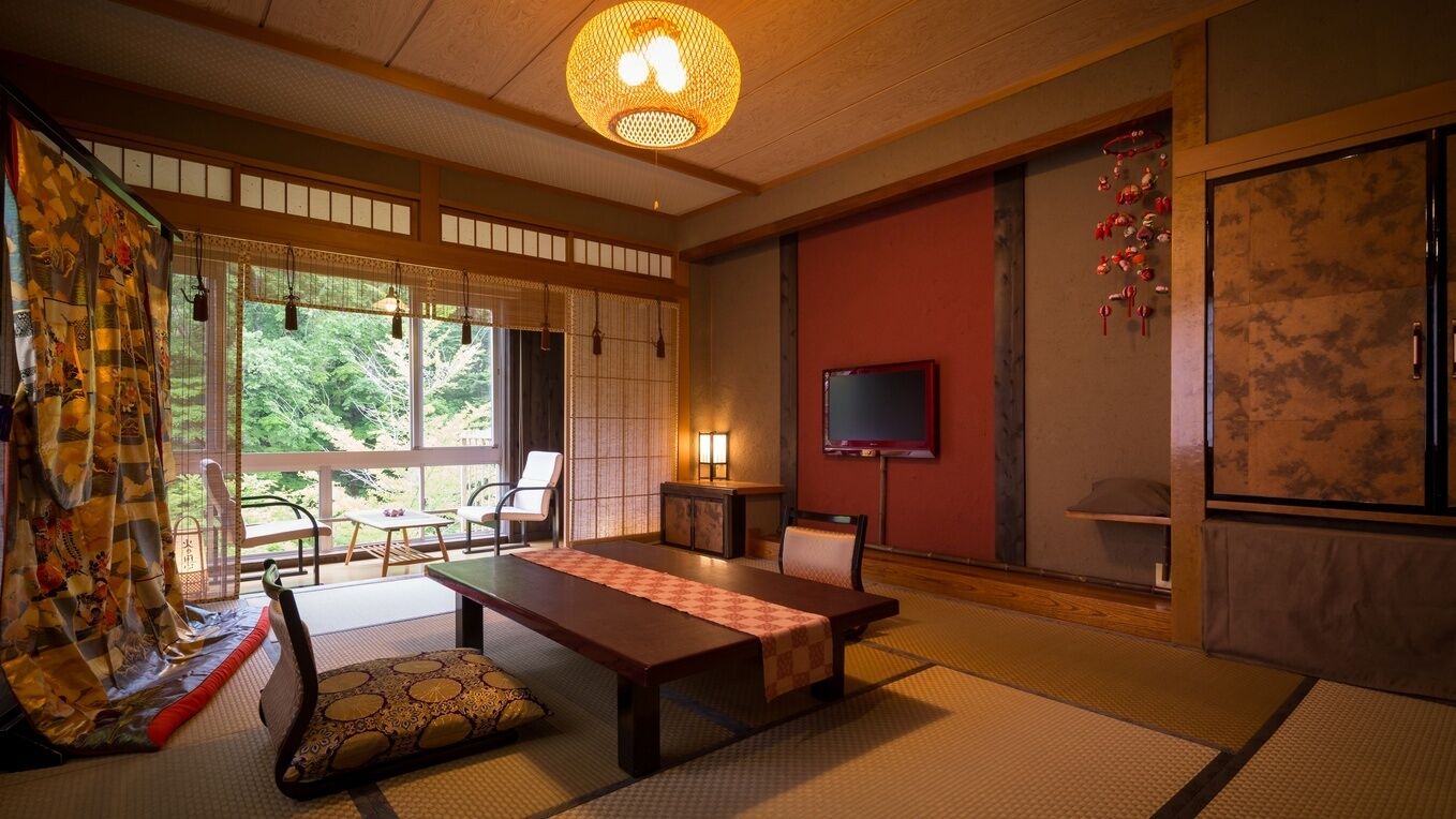 A traditional Japanese room with tatami flooring, a low table, chairs, a television, and a view of greenery outside.