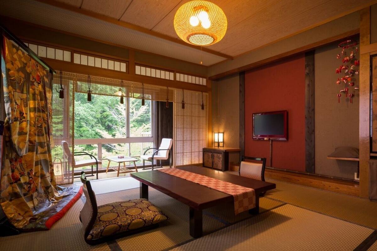 Um quarto japonês tradicional com tatames, uma mesa baixa, cadeiras, uma televisão e vista para o verde lá fora.