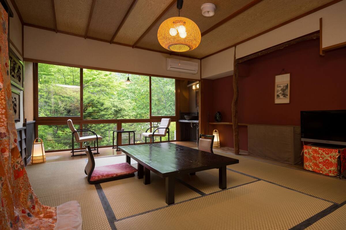 Quarto japonês tradicional com tatames, uma mesa baixa, cadeiras, televisão e uma grande janela com vista para o verde.