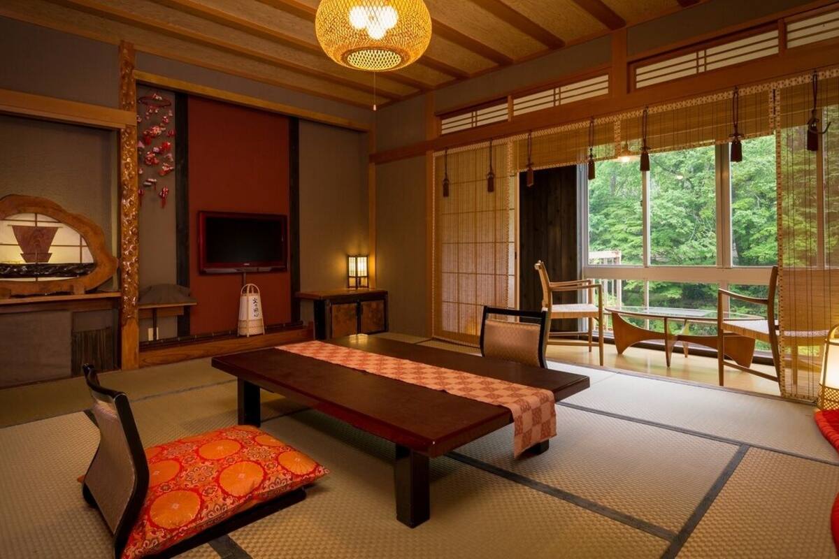 Quarto japonês tradicional com tatames, uma mesa baixa, cadeiras, televisão e vista para o verde através de grandes janelas.